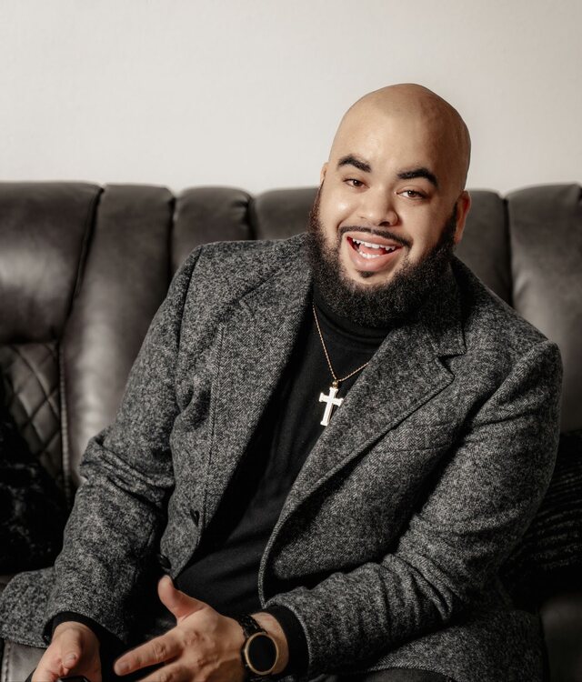 Portrait of Dr. Goodluck Ijezie-Desbois, seated and smiling, wearing a herringbone blazer over a black turtleneck.